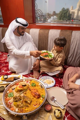 The atmosphere of the blessed month of Ramadan, a breakfast table filled with delicious foods and appetizers, strengthening the bond of fatherhood between the father and his son, an Emirati Gulf Arab man sitting in a traditional ground seating area having breakfast with his son, gestures of happiness and joy.