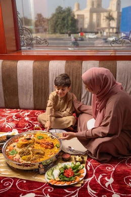 سفرة إفطار مليئة بأصناف من المأكولات والمقبلات الشهية، تقوية رابط الأمومة بين الأم و ابنها ، امراه عربيه خليجيه اماراتية ترتدي العباءة و الحجاب تجلس فى مجلس ارضي شعبي تقوم بتناول الافطار مع ابنها ، الاشارة الي شيء ما، ايماءات السعادة و السرور