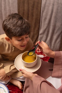 Strengthening the maternal bond between the mother and her son, light meals and healthy food, a close-up shot of the hand of an Emirati Gulf Arab woman serving her son soup, an Emirati Gulf Arab boy eating the soup.