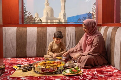 Spending enjoyable times with children, the role and affection of a mother in caring for her children, the concept of a healthy diet, an Emirati Gulf Arab woman wearing an abaya and hijab sitting in a traditional floor seating area with her son as they have Iftar during Ramadan.