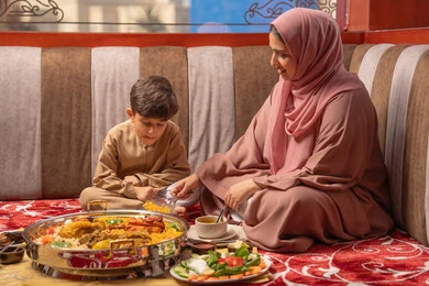 دور و حنان الأم في رعايه أبنائها، مفهوم النظام الغذائي الصحي، قضاء أوقات ممتعة مع الأبناء،  امراه عربيه خليجيه اماراتية ترتدي العباءة و الحجاب تجلس فى مجلس ارضي شعبي برفقة ابنها يقومان بتناول افطار رمضان
