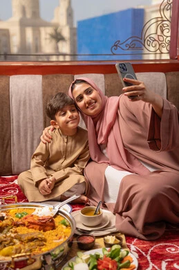 Using a mobile phone to take selfies, the concept of a happy family, an Arab Gulf Emirati woman wearing an abaya and hijab sitting in a traditional floor seating area with her son, a dining table filled with various types of food, gestures of happiness and joy.