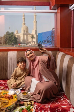 Taking a selfie with a mobile phone, the concept of a happy family, an Arab Gulf Emirati woman wearing an abaya and hijab sitting in a traditional floor seating area with her son, a dining table filled with various types of food, gestures of happiness and joy.