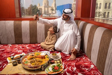 التقاط صور السيلفي عبر الهاتف المحمول، مفهوم الأسرة السعيدة، رجل عربي خليجي اماراتي يرتدي الكندورة و الغتره يجلس فى مجلس ارضي شعبي برفقة ابنه ، مائدة طعام مليئة بانواع مختلفة من المأكولات، ايماءات السعادة و السرور