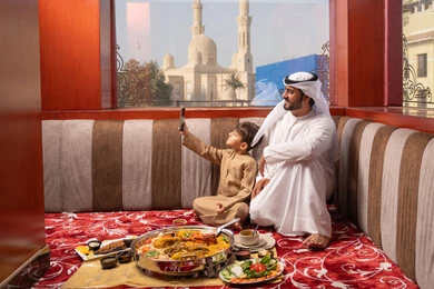 مفهوم الأسرة السعيدة، رجل عربي خليجي اماراتي يرتدي الكندورة و الغتره يجلس فى مجلس ارضي شعبي برفقة  ابنه ، التقاط صور السيلفي عبر الهاتف المحمول،  مائدة طعام مليئة بانواع مختلفة من المأكولات، ايماءات السعادة و السرور