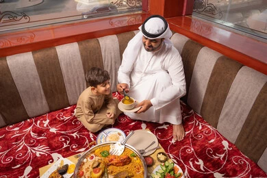 المأكولات الشرقية الشهية، مفهوم الترابط الأسري، اجتماع الأسرة على السفرة, رجل عربي خليجي اماراتي يرتدي الكندورة و الغتره يجلس فى مجلس شعبي ارضي برفقة ابنه يقوم بتناول الشوربة, الأجواء الرمضانية العائلية السعيدة