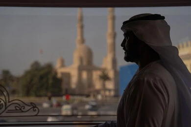 ايماءات الاندماج و التأمل، روحانيات ومعمار إسلامي، صورة ظلية من الخلف لرجل عربي خليجي إماراتي يرتدي الكندورة و الغتره يقف خلف النافذة يشاهد المسجد