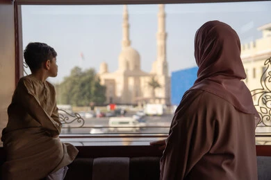 قضاء أوقات مميزة مع الأسرة، الاعتناء بالأطفال والاهتمام بهم, مشاعر الأمومة و العطف، صورة من الخلف لامراه عربيه خليجيه اماراتية ترتدي العباءة و الحجاب تقف بجانب ابنها يتأملان المسجد والمعالم العمرانية من خلف النافذة