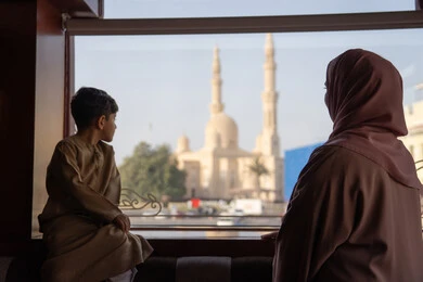 الاعتناء بالأطفال والاهتمام بهم, مشاعر الأمومة و العطف،  قضاء أوقات مميزة مع الأسرة، صورة من الخلف لامراه عربيه خليجيه اماراتية ترتدي العباءة و الحجاب تقف بجانب ابنها يتأملان المسجد والمعالم العمرانية من خلف النافذة