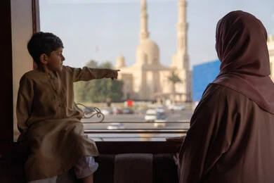 الاشارة الي شيء ما، قضاء أوقات مميزة مع الأسرة،  صورة من الخلف لامراه عربيه خليجيه اماراتية ترتدي العباءة و الحجاب تقف بجانب ابنها يتأملان المسجد والمعالم العمرانية من خلف النافذة