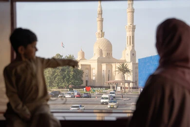 Spending special times with family, pointing to something, a picture from behind of an Arab Gulf Emirati woman wearing an abaya and hijab standing next to her son as they contemplate the mosque and architectural landmarks from behind the window.