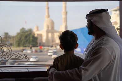 ايماءات الحب والحنان، قضاء وقت عائلي ممتع، صورة من الخلف لرجل عربي خليجي اماراتي يرتدي الكندورة و الغتره يقف بجانب ابنه يتأملان المسجد والمعالم العمرانية من خلف النافذة