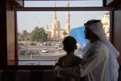 قضاء وقت عائلي ممتع، صورة من الخلف لرجل عربي خليجي اماراتي يرتدي الكندورة و الغتره يقف بجانب ابنه يتأملان المسجد والمعالم العمرانية من خلف النافذة