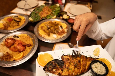 سفرة رمضانية مليئه بالمأكولات الذيذة ، فنون التقديم في مطاعم ومطابخ الامارات العربية، صورة مقربة ليد رجل عربي خليجي اماراتي يمسك بيده طبق من السمك المشوي