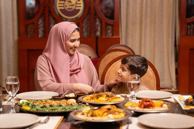 Delicious foods and appetizers, family gatherings around the dining table, a family atmosphere filled with love, discussions and family dialogues, a dining table filled with a variety of Ramadan meals, Islamic holidays and occasions, an Emirati Gulf Arab mother wearing an abaya and hijab having breakfast with her son, the month of goodness and blessings.
