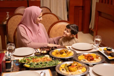 The family gathers around the dining table, a family atmosphere filled with love, discussions and family dialogues, a table full of diverse Ramadan meals, Islamic holidays and occasions, an Emirati Gulf Arab mother wearing an abaya and hijab having breakfast with her son, delicious dishes and appetizers, the month of goodness and blessings.