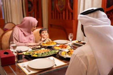 A breakfast table filled with a variety of Emirati Ramadan dishes, Islamic and religious occasions, gestures of happiness and joy, an Arab Gulf Emirati woman wearing an abaya and hijab sits at the table having breakfast with her husband and son.