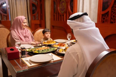 إفطار رمضاني غني بالمأكولات العربية الإماراتية اللذيذة، المناسبات الاسلامية و الدينية، ايماءات السعادة و السرور،  امراه عربيه خليجيه اماراتية ترتدي العباءة و الحجاب تجلس على الطاولة تقوم بتناول الافطار مع زوجها و ابنها