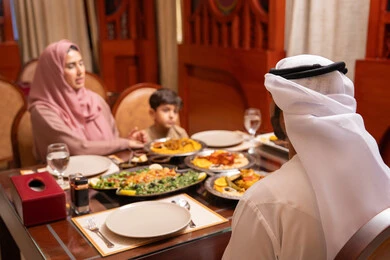وجبة إفطار متنوعة من الأطباق الإماراتية الرمضانية على الطاولة، المناسبات الاسلامية و الدينية، امراه عربيه خليجيه اماراتية ترتدي العباءة و الحجاب تجلس على الطاولة تقوم بتناول الافطار مع زوجها و ابنها