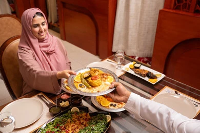Gestures of happiness and joy, a Ramadan iftar table with a variety of delicious Emirati dishes, an Arab Gulf Emirati woman wearing an abaya and hijab sitting at the table having iftar with her husband, Islamic and religious occasions.