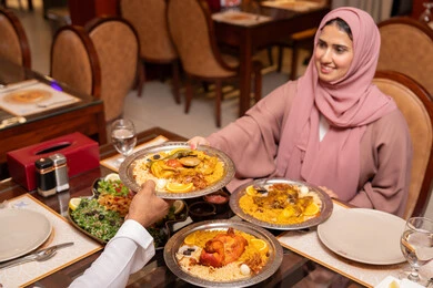 A Ramadan iftar table with a variety of delicious Emirati dishes, an Arab Gulf Emirati woman wearing an abaya and hijab sits at the table having iftar with her husband, gestures of happiness and joy, Islamic and religious occasions.