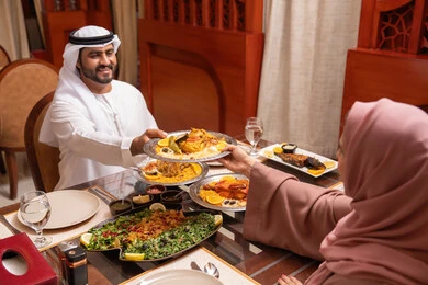 إفطار يحتوي على تشكيلة من المأكولات العربية الإماراتية الرمضانية،رجل عربي خليجي اماراتي يرتدي الكندورة و الغتره يجلس على الطاولة يقوم بتناول الافطار مع زوجته ، ايماءات السعادة و السرور، المناسبات الاسلامية و الدينية