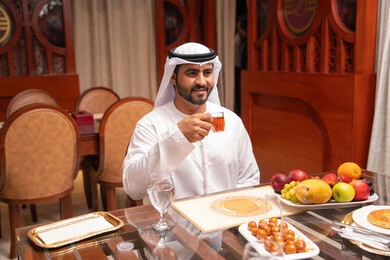 Enjoying hot drinks inside a café, indulging in recreational activities, spending enjoyable times, expressions of joy and happiness, an Emirati Gulf Arab man wearing a kandura and ghutrah sitting at a table holding a cup of tea, restaurants and cafes in the United Arab Emirates.