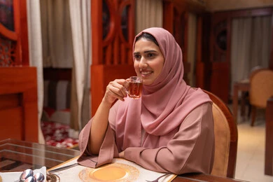 الاستمتاع بشرب المشروبات الساخنة في احدى الكافيهات، الاستمتاع بالأنشطة الترفيهية، قضاء أوقات ممتعة في المقهى، تعابير السرور والسعادة، امراه عربيه خليجيه اماراتية ترتدي العباءة و الحجاب تمسك بيديها كوب من الشاي , مطاعم و كافيهات في الإمارات العربية المتحدة