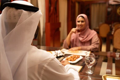 الاستمتاع بالأنشطة الترفيهية، قضاء أوقات ممتعة في المقهى، تعابير السرور والسعادة، وصفات وأطباق حلويات،صورة من الخلف لرجل عربي خليجي اماراتي يرتدي الكندورة والغترة البيضاء يجلس على الطاولة يقوم بتناول كوب من الشاي برفقة زوجته, مطاعم و كافيهات في الإمارات العربية المتحدة