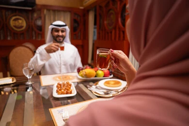 قضاء أوقات ممتعة في المقهى، تعابير السرور والسعادة، وصفات وأطباق حلويات، رجل عربي خليجي اماراتي يرتدي الكندورة والغترة البيضاء يجلس على الطاولة يقوم بتناول كوب من الشاي برفقة زوجته ، الاستمتاع بالأنشطة الترفيهية, مطاعم و كافيهات في الإمارات العربية المتحدة