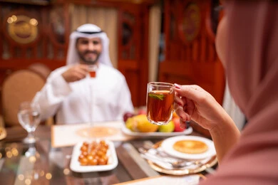 Recipes and dishes of desserts, expressions of joy and happiness, spending enjoyable times in the café, a close-up image from behind of an Emirati Gulf Arab woman wearing a hijab sitting at the table enjoying a cup of tea with her husband, enjoying recreational activities, restaurants and cafes in the United Arab Emirates.