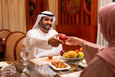 Taking care of fitness and physical health through healthy nutrition, the holy month and the spiritual Ramadan atmosphere, spending enjoyable times with family outdoors, an Arab Gulf Emirati man wearing the kandura and ghutrah is giving his wife an apple with gestures of happiness and joy.