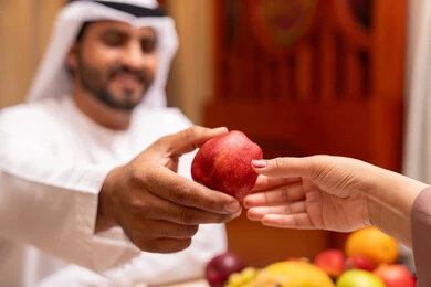 Caring about fitness and health through healthy food, the holy month and the spiritual Ramadan atmosphere, spending enjoyable times with family outdoors, a close-up image of an Emirati Gulf Arab man wearing a kandura and ghutrah giving his wife an apple with gestures of happiness and joy.