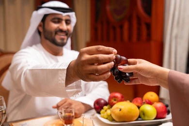 الشهر الكريم والأجواء الرمضانية الروحانية، الحفاظ على الرشاقة والصحه البدنية من خلال تناول الطعام المفيد، قضاء أوقات ممتعه مع العائله في الخارج، رجل عربي خليجي اماراتي يرتدي الكندورة و الغتره يقوم باعطاء زوجته العنب بايماءات السعادة و السرور