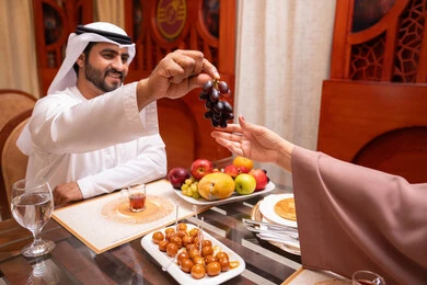 Maintaining fitness and physical health through eating beneficial food, the holy month and the spiritual Ramadan atmosphere, spending enjoyable times with family outdoors, an Arab Gulf Emirati man wearing a kandura and ghutrah is giving his wife grapes with gestures of happiness and joy.