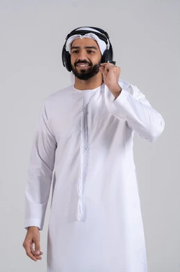 Assisting customers through communication to resolve issues, answering questions and inquiries, integrating advanced technologies with work, looking at the camera with expressions of happiness and joy, a portrait of a young Arab Gulf Emirati man wearing a kandura and ghutrah, wearing headphones, working in the customer service department, with a white background.