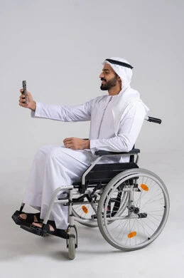 The concept of elegance, masculinity, and attention to appearance, preserving happy moments through photos, a side portrait of an Arab Gulf Emirati man with special needs wearing a kandura and a white ghutrah sitting in a wheelchair holding a mobile phone and taking a selfie, gestures of self-confidence, white background.