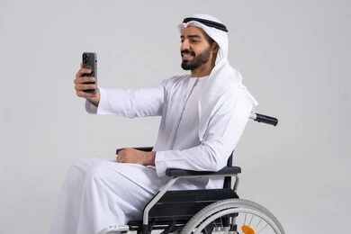 Preserving happy moments through photos, the concept of elegance, masculinity, and attention to appearance, a side portrait of an Arab Gulf Emirati man with special needs wearing a kandura and a white ghutrah sitting in a wheelchair holding a mobile phone and taking a selfie, gestures of self-confidence, white background.
