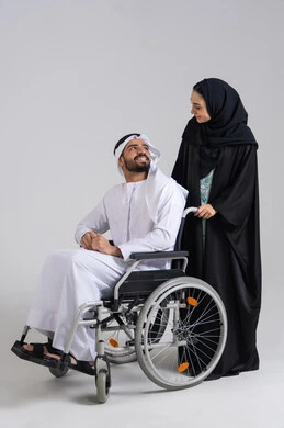 An Arab Gulf Emirati woman helps her husband with mobility and movement, the loyalty and mutual affection between the spouses, caring for people with determination and securing their needs, the lifestyle of individuals with mobility disabilities, providing tools and equipment to assist patients, a Gulf man with special needs sitting in a wheelchair looking at his wife, white background.