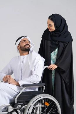 الوفاء والحنان المتبادل بين الزوجين، العنايه بأصحاب الهمم وتأمين احتياجاتهم ، نمط حياة ذوي الإعاقة الحركية ، تأمين أدوات ومعدات لمساعدة المرضي ، رجل خليجي من ذوي الاحتياجات الخاصه يجلس على كرسي متحرك ينظر الى زوجته، امراة عربيه خليجيه اماراتية تساعد زوجها على التنقل والحركة ، خلفية بيضاء