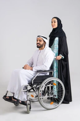 نمط حياة ذوي الإعاقة الحركية ،امراة عربيه خليجيه اماراتية تساعد زوجها على التنقل والحركة، الوفاء والحنان المتبادل بين الزوجين، العنايه بأصحاب الهمم وتأمين احتياجاتهم ، تأمين أدوات ومعدات لمساعدة المرضي ، رجل خليجي من ذوي الاحتياجات الخاصه يجلس على كرسي متحرك ، خلفية بيضاء