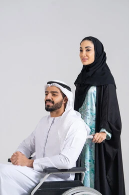 Providing tools and equipment to assist patients, the lifestyle of individuals with mobility disabilities, an Arab Gulf Emirati woman helping her husband to move and navigate, the mutual loyalty and affection between spouses, caring for people of determination and securing their needs, a Gulf man with special needs sitting in a wheelchair, white background.