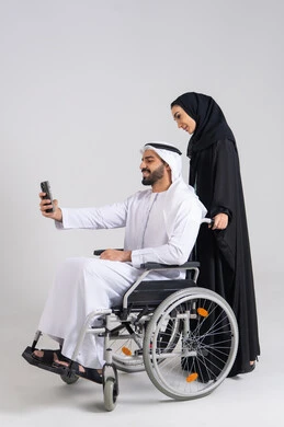 Taking commemorative selfie photos using a mobile phone, focusing on the psychological and physical aspects of the disabled, caring for patients and the disabled, securing equipment that aids in movement and mobility, a portrait of an Emirati Gulf Arab wife wearing an abaya and hijab helping her husband who has special needs, with a white background.
