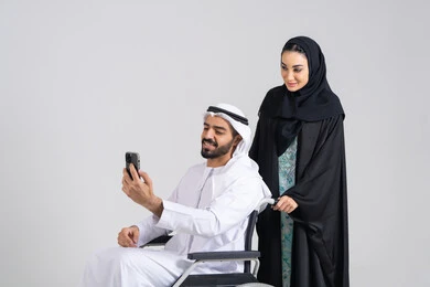Caring for the psychological and physical aspects of the disabled, taking care of patients and the disabled, providing equipment to assist with movement and mobility, taking commemorative selfies using a mobile phone, a portrait of an Emirati Gulf Arab wife wearing an abaya and hijab helping her husband with special needs, white background.
