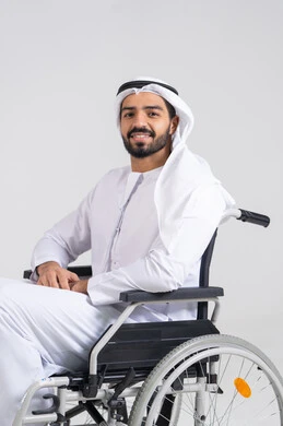النظرالى الكاميرا بايماءات السعادة و السرور، توفير اجهزة ومعدات تساعد بالحركة والتنقل، تحسين حياة ذوي الاعاقه الحركيه، بورترية لرجل عربي خليجي اماراتي من ذوى الاحتياجات الخاصة يرتدي الكندورة و الغترة البيضاء يجلس على كرسي متحرك، خلفيه بيضاء
