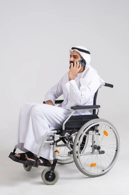 التواصل مع الاهل والاصدقاء، اجراء مكالمات هاتفية، ايماءات السرور والسعادة ،استخدام الاجهزة التقنية الحديثة، بورتريه من الجانب  لرجل عربي خليجي اماراتي مقعد يرتدي الكندورة و الغتره يجلس على كرسي متحرك يحمل بيده الهاتف المحمول، خلفية بيضاء