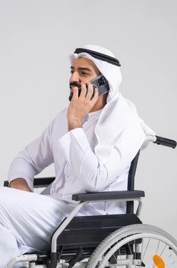 Making phone calls, gestures of joy and happiness, using modern technological devices, communicating with family and friends, a side portrait of an Emirati Gulf Arab man sitting in a wheelchair wearing a kandura and ghutrah, holding a mobile phone in his hand, with a white background.
