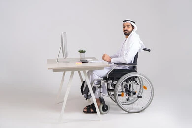 ايماءات السعادة و السرور، إنجاز مهام العمل، إدارة شؤون الأعمال وتنظيمها عبر الاجهزة التقنية، دمج العمل مع التقنيات والتكنولوجيا، مهن ووظائف ادارية مكتبية, صورة من الجانب لرجل عربي خليجي اماراتي يرتدي الكندورة و الغترة يجلس على كرسي متحرك يعمل على الحاسوب داخل المكتب