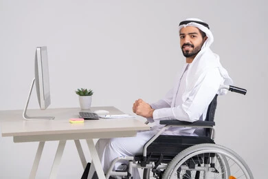 النظر الى الكاميرا بتعابير السعادة و السرور، إنجاز مهام العمل، إدارة شؤون الأعمال وتنظيمها عبر الاجهزة التقنية، دمج العمل مع التقنيات والتكنولوجيا، مهن ووظائف ادارية مكتبية, صورة من الجانب لرجل عربي خليجي اماراتي يرتدي الكندورة و الغترة يجلس على كرسي متحرك يعمل على الحاسوب داخل المكتب