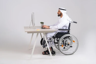 Completing work tasks, managing and organizing business affairs through technical devices, integrating work with technologies and technology, administrative and office job professions, a side view of an Arab Gulf Emirati man wearing a kandura and ghutrah sitting in a wheelchair working on a computer inside the office.